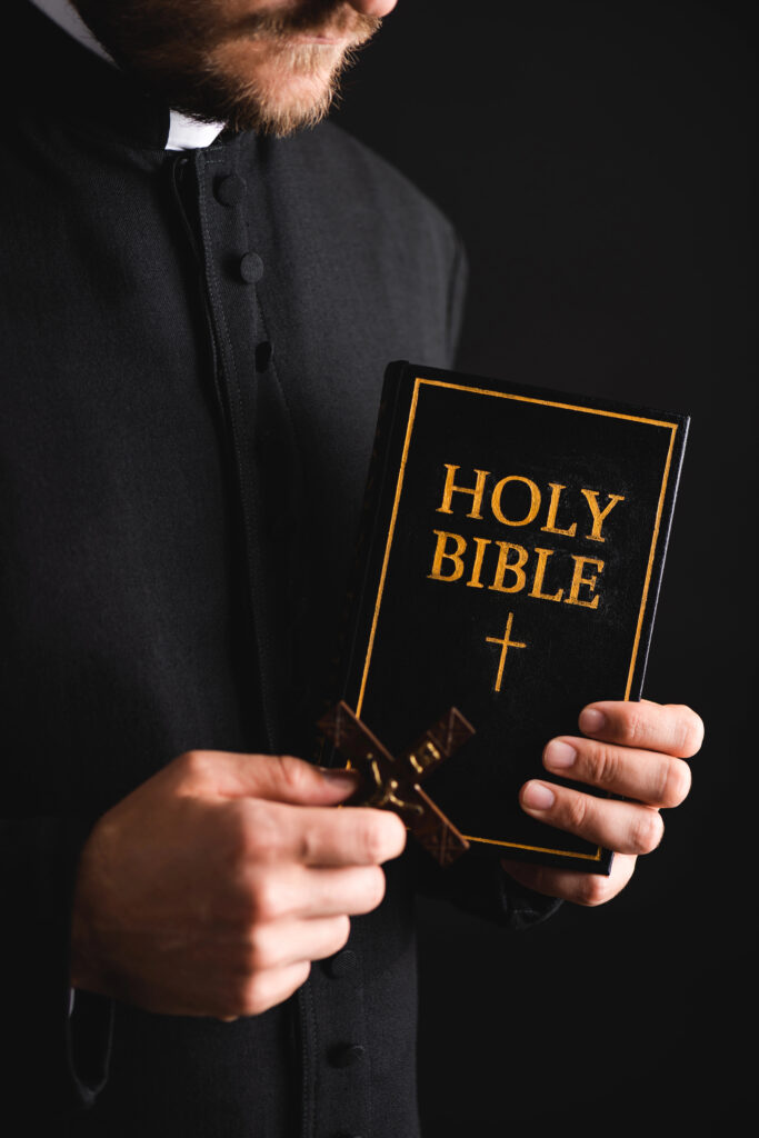 Depositphotos_391958882_XL Cropped view of bearded priest holding cross and holy bible isolated on black Author HayDimitriy